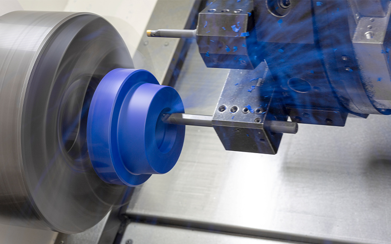Ein blaues Kunststoff-Bauteil eingespannt in eine CNC-Maschine wird gedreht. Dabei fliegen Kunststoff-Späne.