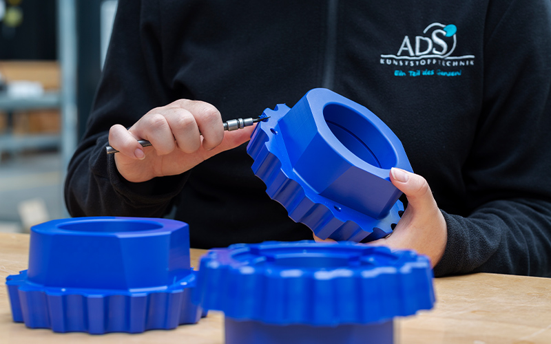 Ein Mitarbeiter von ADS Kunststofftechnik hält ein blaues Kunststoffbauteil in den Händen und entgratet es mit einem schmalen Werkzeug.