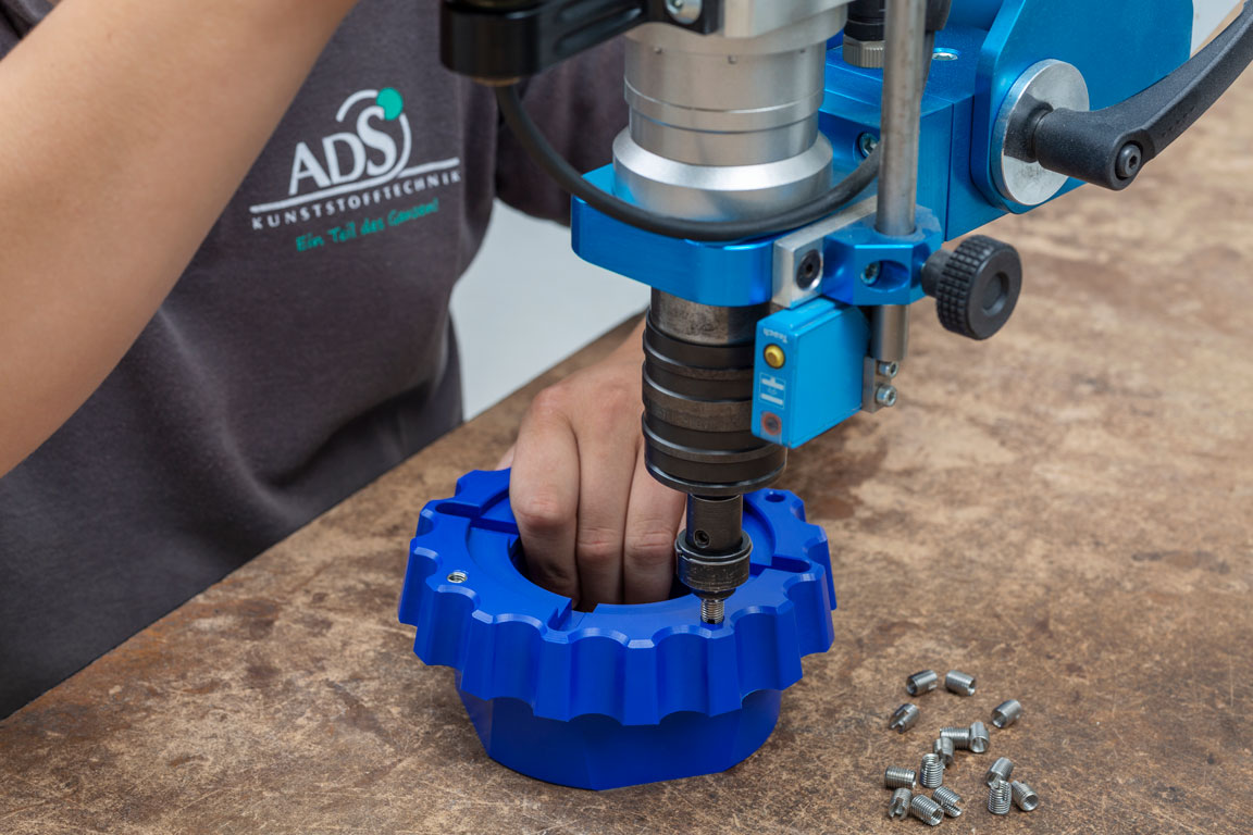 Ein Mitarbeiter der ADS Kunststofftechnik hält mit einer Hand ein blaues Kunststoffbauteil und dreht mit Hilfe einer Maschine Gewindebuchsen in das Kunststoffteil ein.