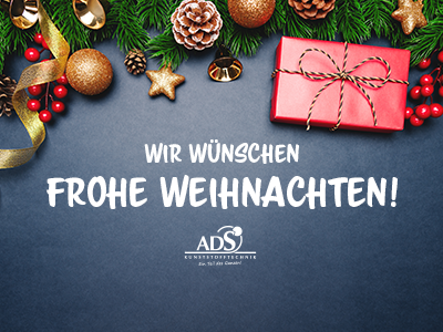 Auf einem grauen Tisch liegt ein rotes Geschenk, Tannenzapfen und -zweige. Auf dem Tisch steht "Wir wünschen Frohe Weihnachten".