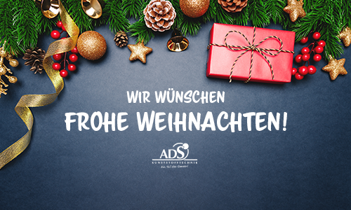 Auf einem grauen Tisch liegt ein rotes Geschenk, Tannenzapfen und -zweige. Auf dem Tisch steht "Wir wünschen Frohe Weihnachten".